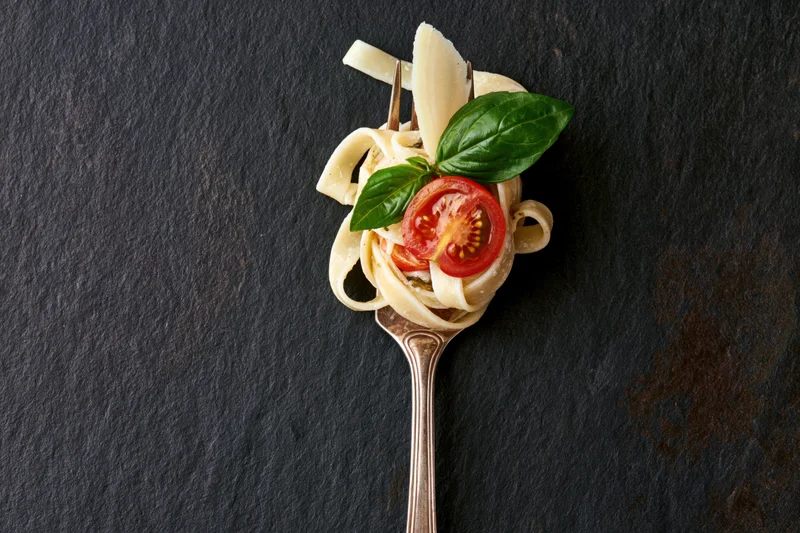 Vintage silver fork twirled with fettuccine pasta topped with tomato, basil, and parmesan cheese on dark slate surface