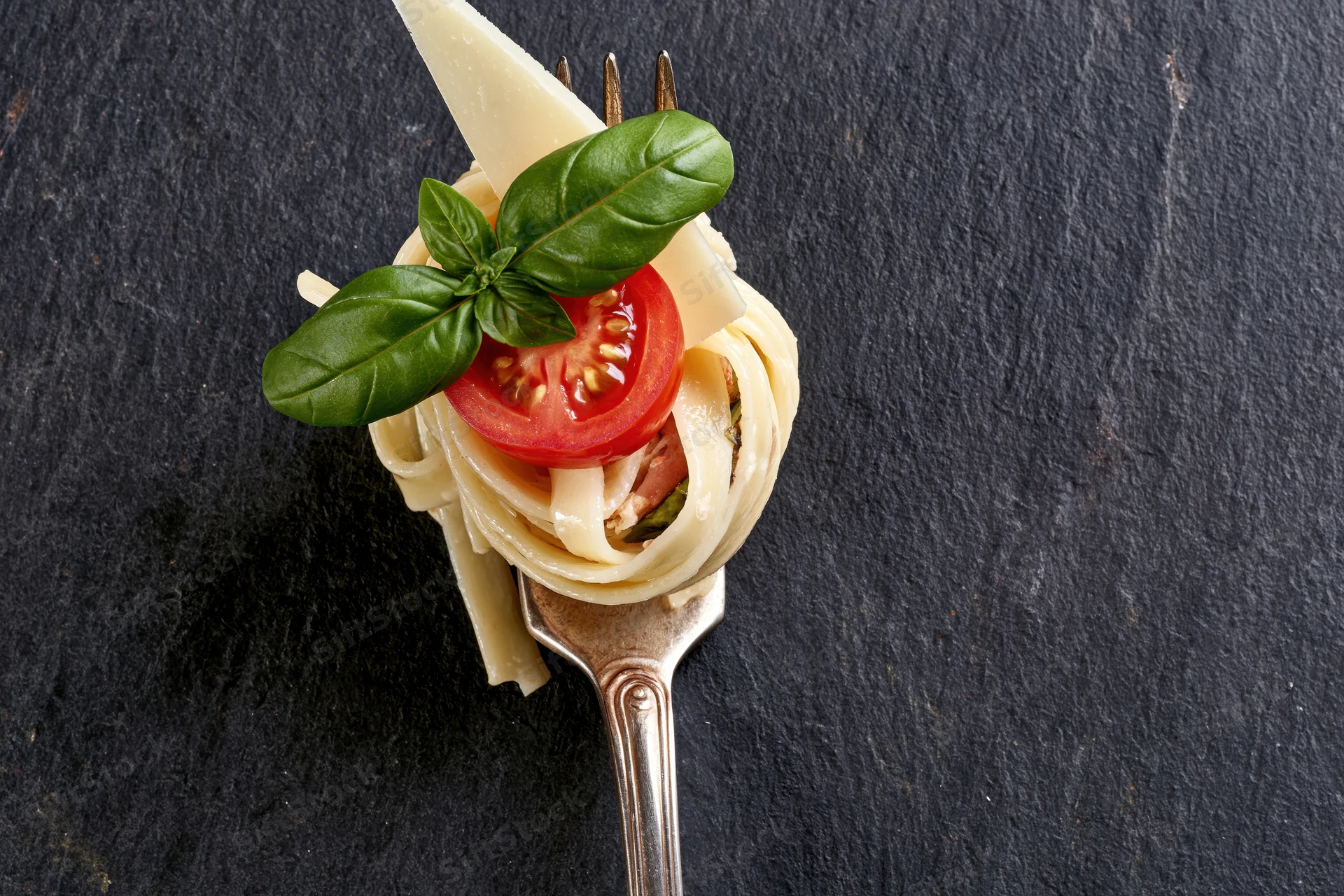 Vintage silver fork twirled with fettuccine pasta topped with tomato, basil, and parmesan cheese on black slate surface