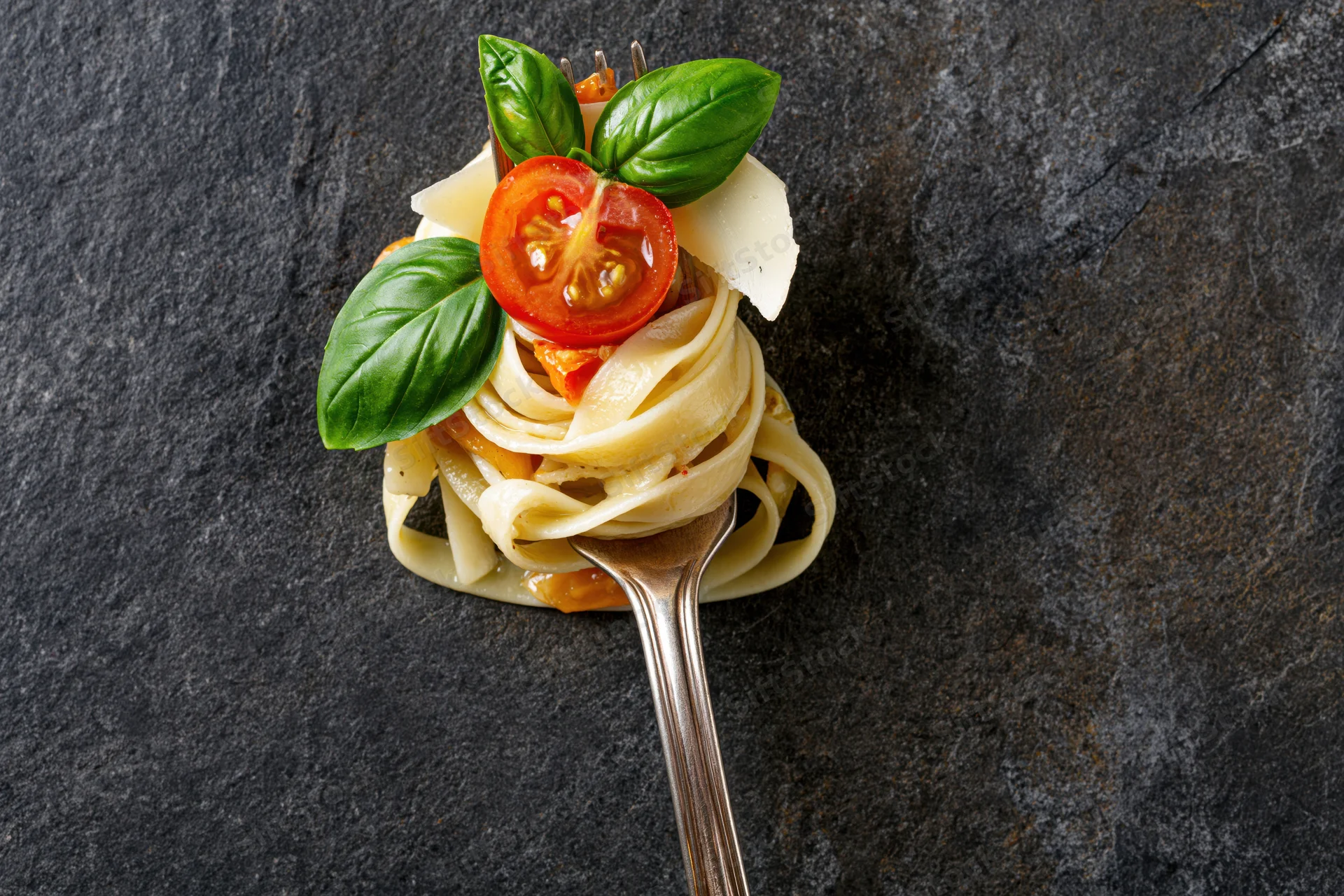 Fettuccine pasta twirled on a vintage silver fork topped with tomato, basil and cheese on dark slate surface