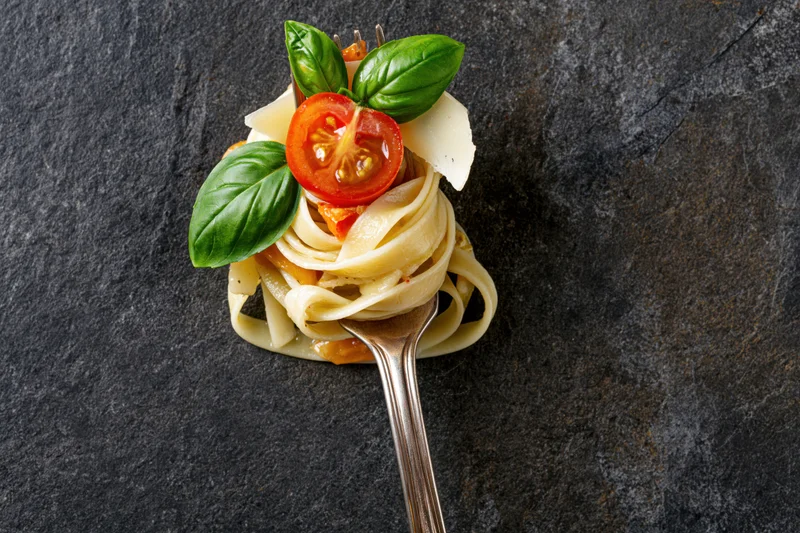 Fettuccine pasta twirled on a vintage silver fork topped with tomato, basil and cheese on dark slate surface