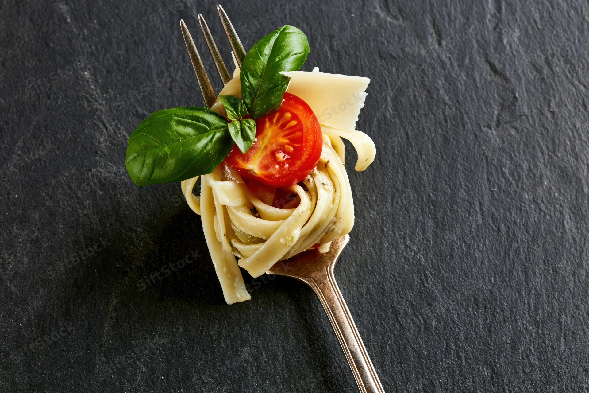 Vintage silver fork twirled with fettuccine pasta topped with basil, tomato, and parmesan cheese on dark slate surface