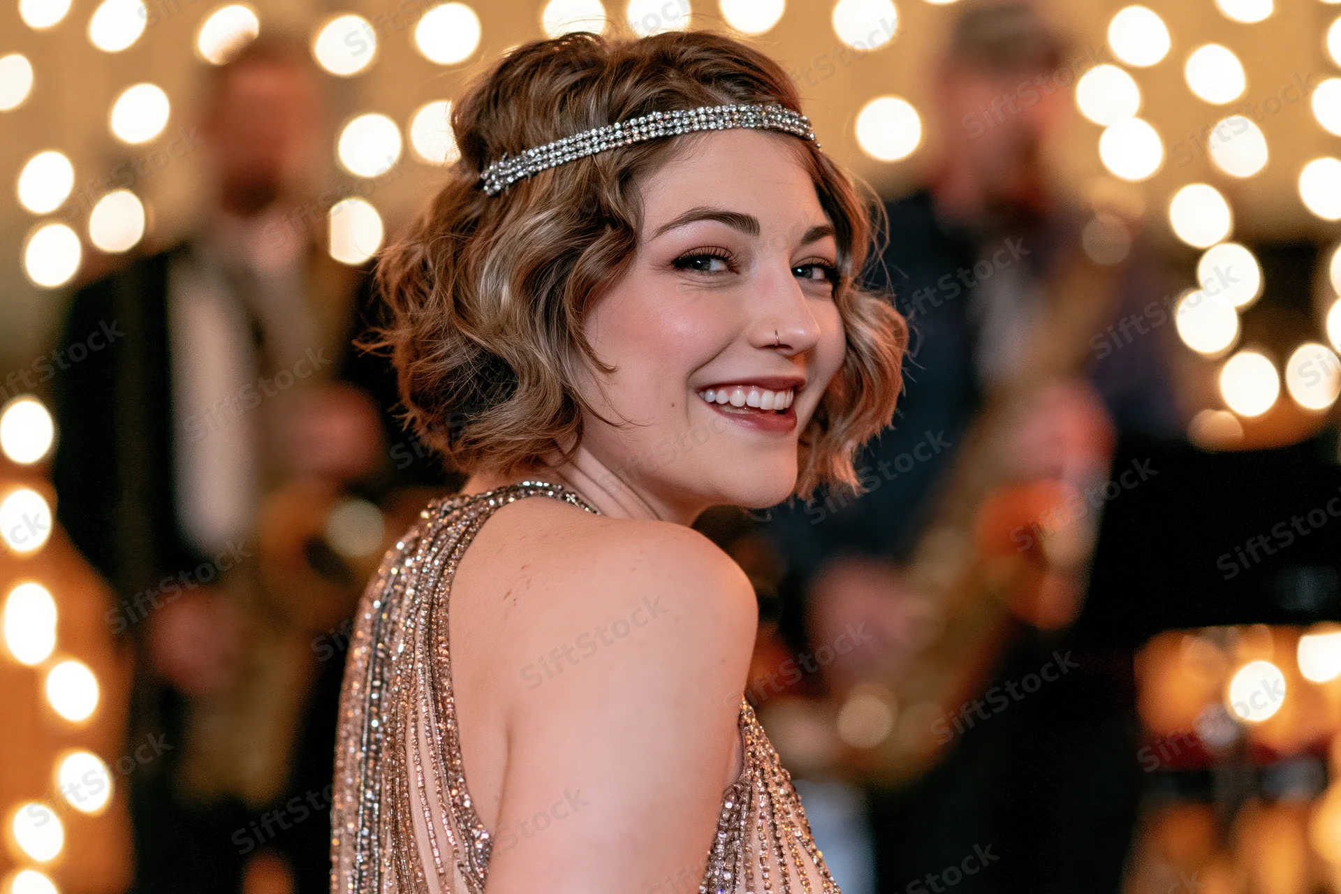 Young woman in 1920s-style beaded halter gown smiling over shoulder at party with musicians and golden lights in background