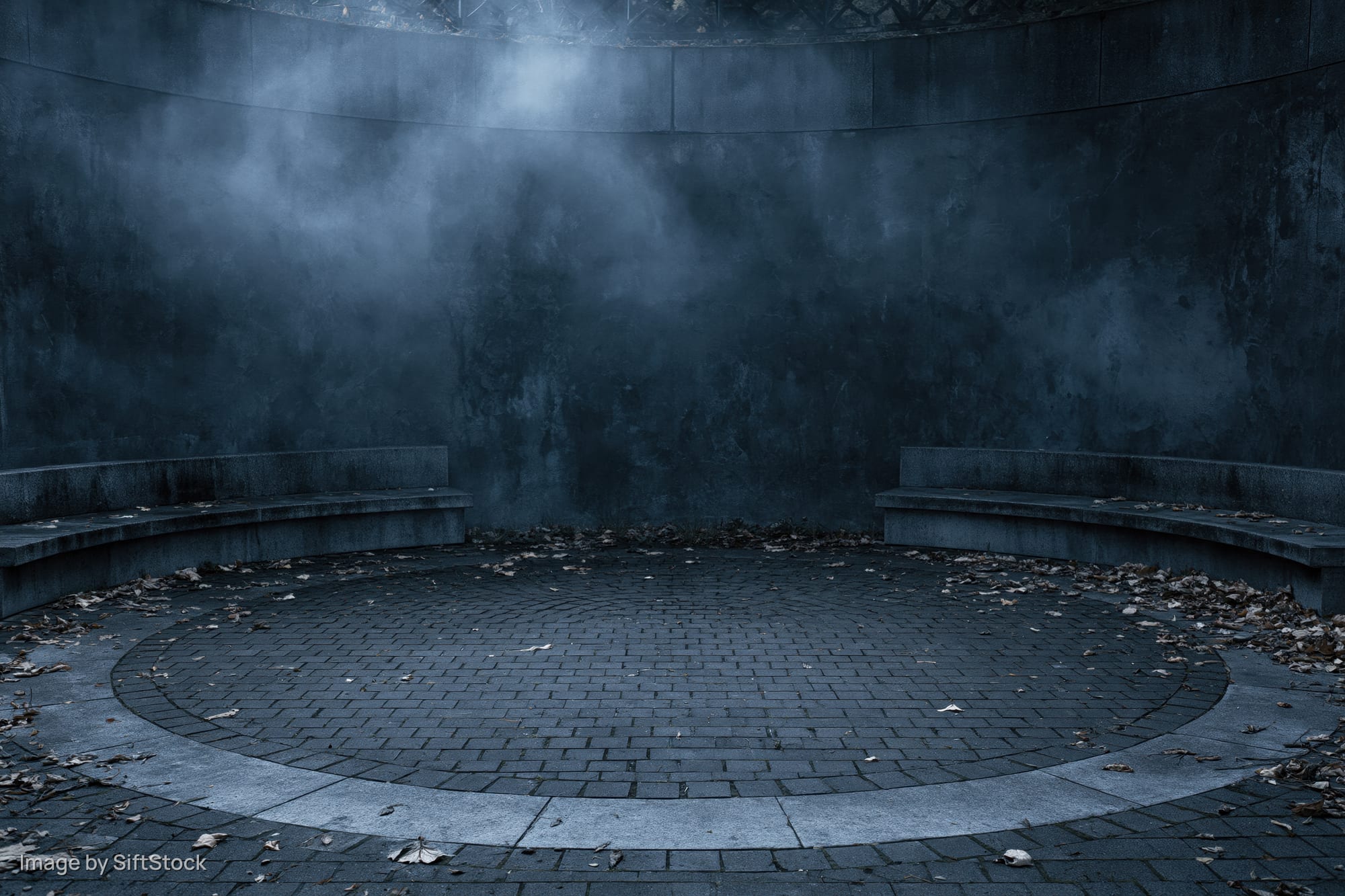 Circular stone courtyard at night with fog and scattered leaves, surrounded by weathered walls and benches
