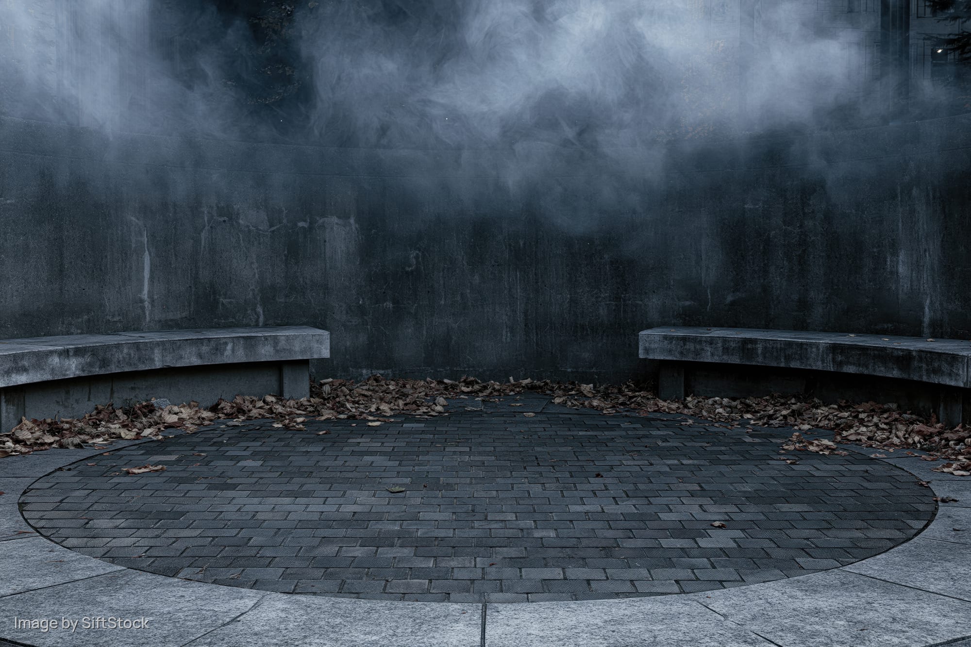 Circular stone courtyard at night with smoke rising between two benches, surrounded by fallen leaves and a weathered concrete wall