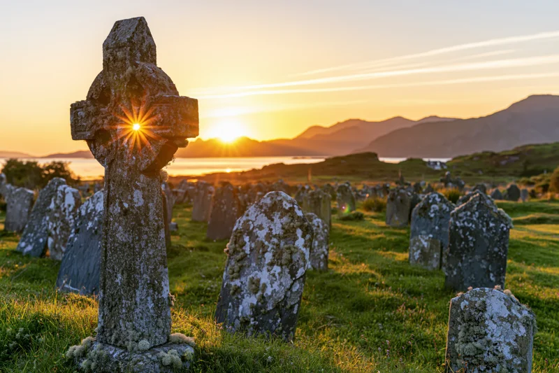 Ancient celtic cross gravestone in a scottish highland cemetery at sunset with mountains and water in the background