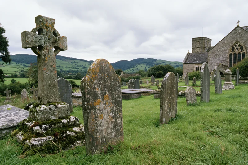 Weathered stone cross and gravestones in a rural cemetery with a church and rolling hills in the background
