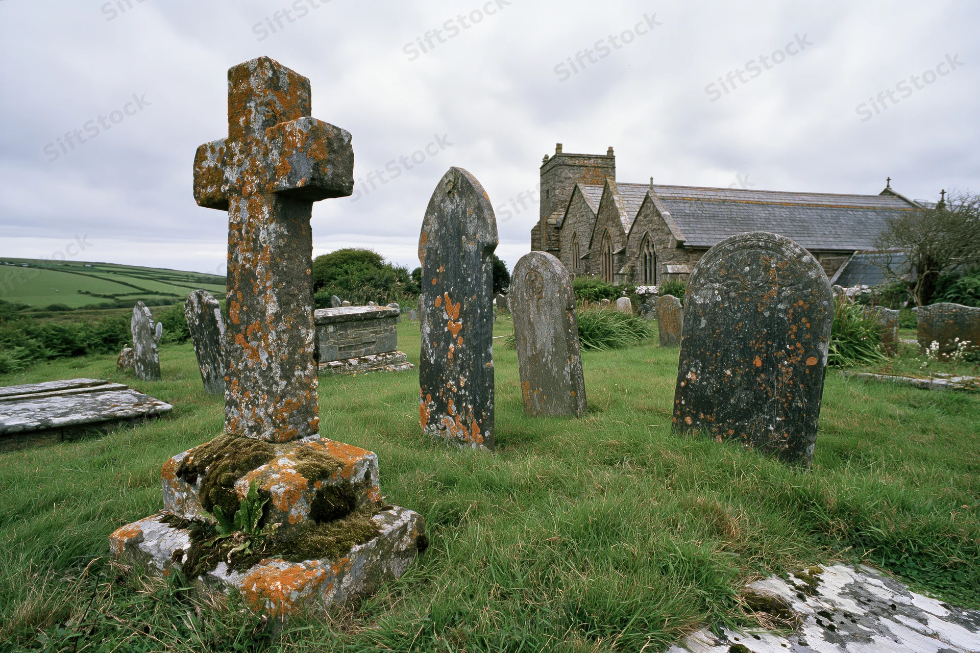 Weathered stone cross and gravestones in a rural graveyard with a church in the background
