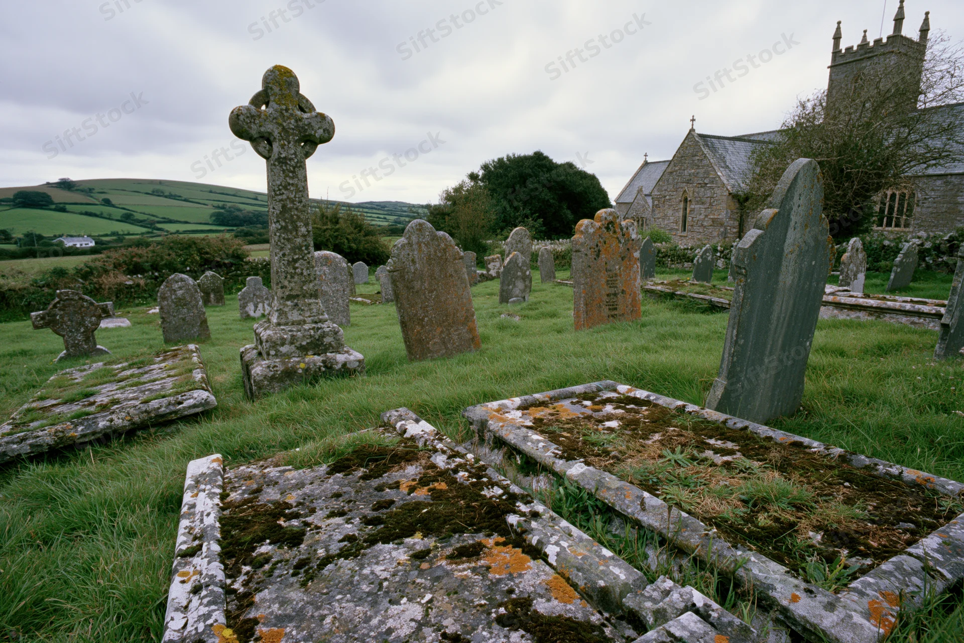 Weathered gravestones and a stone cross in a rural churchyard with rolling hills in the background
