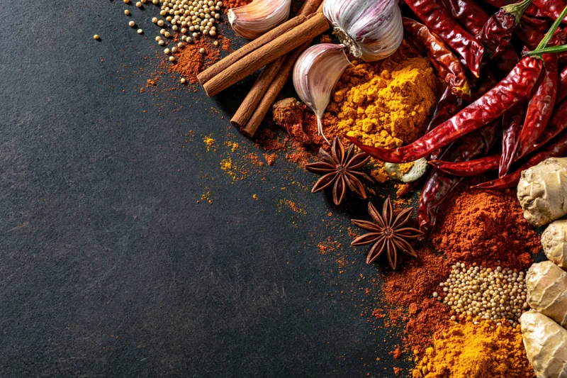 Assorted whole and ground spices arranged along the right side of a dark slate surface with copyspace on the left