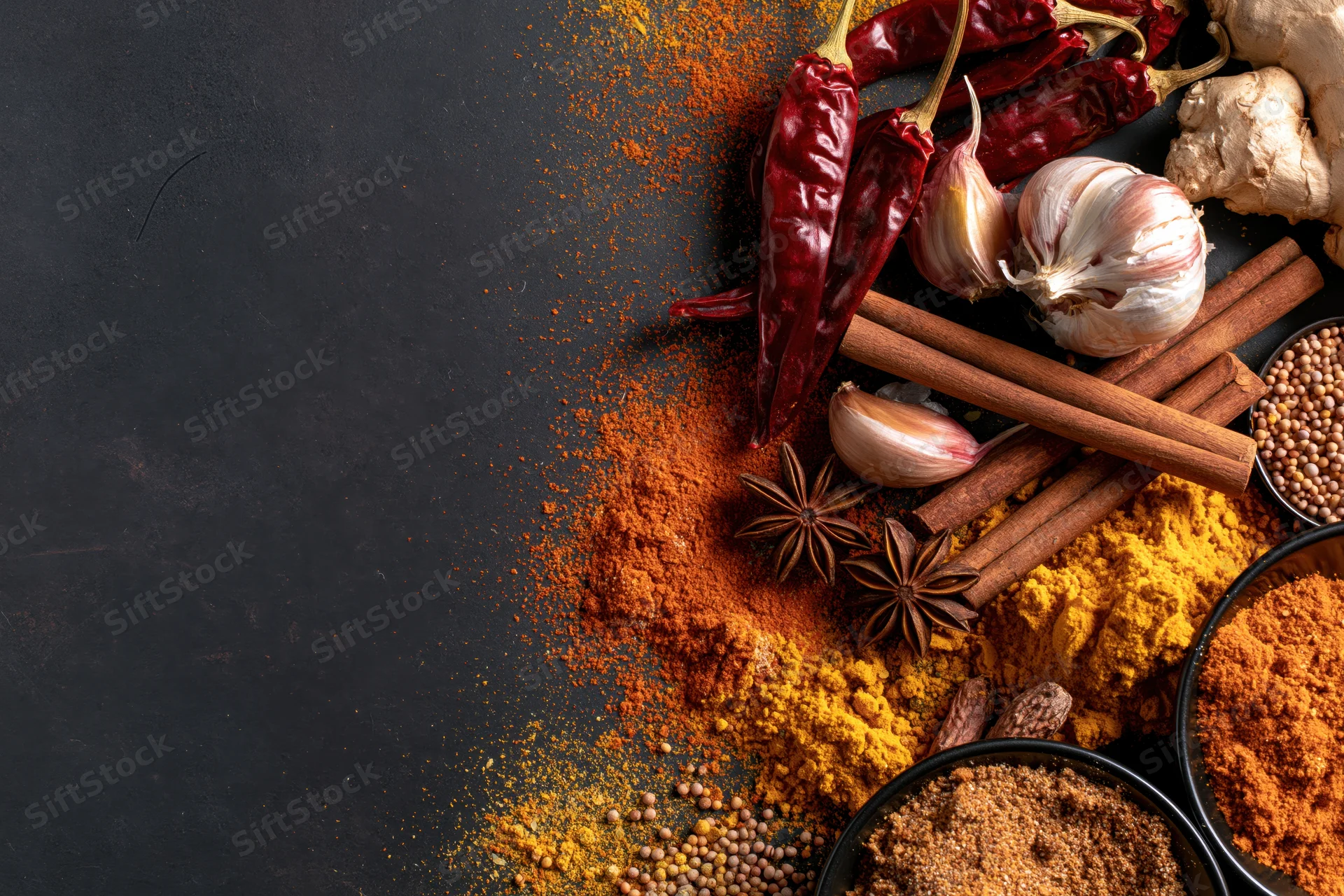 Assorted whole and ground spices arranged along the right side of a dark surface with copyspace on the left