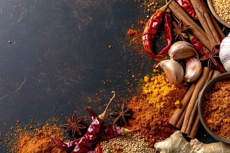 Assorted whole and ground spices arranged along the right side of a dark textured surface with copyspace on the left