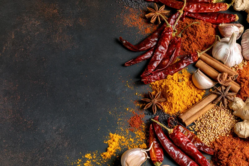 Assorted whole and ground spices arranged along the right side of a dark textured surface with copyspace on the left