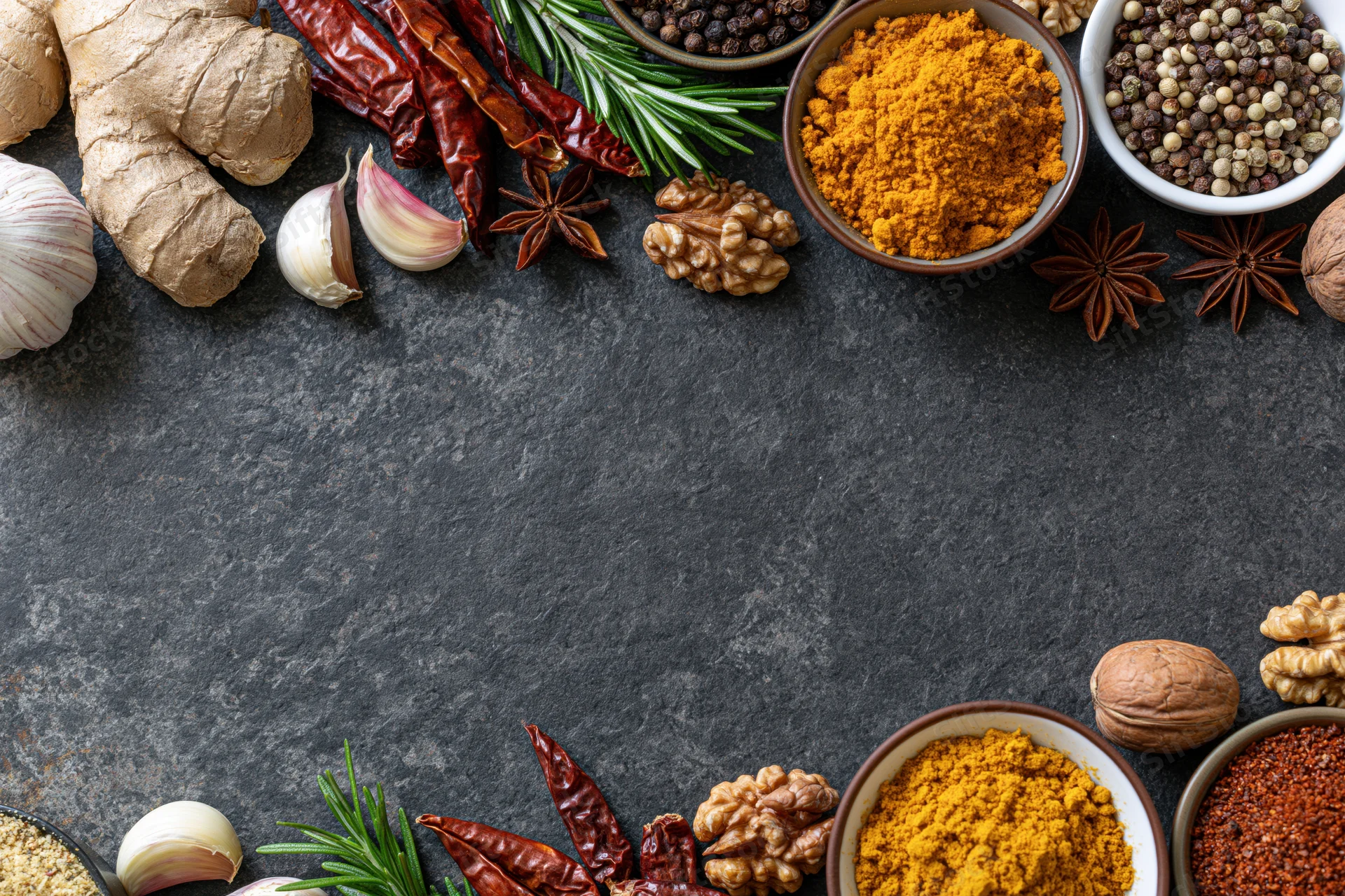 Assorted spices and herbs arranged along the top and bottom edges of a dark surface with copyspace in the center