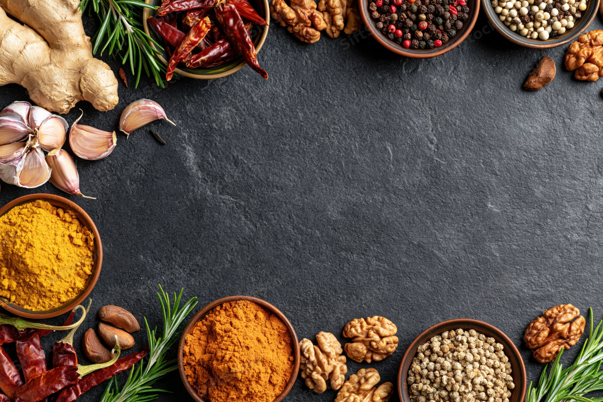 Assorted whole and ground spices arranged along the top and bottom edges of a dark slate surface with copyspace in the center