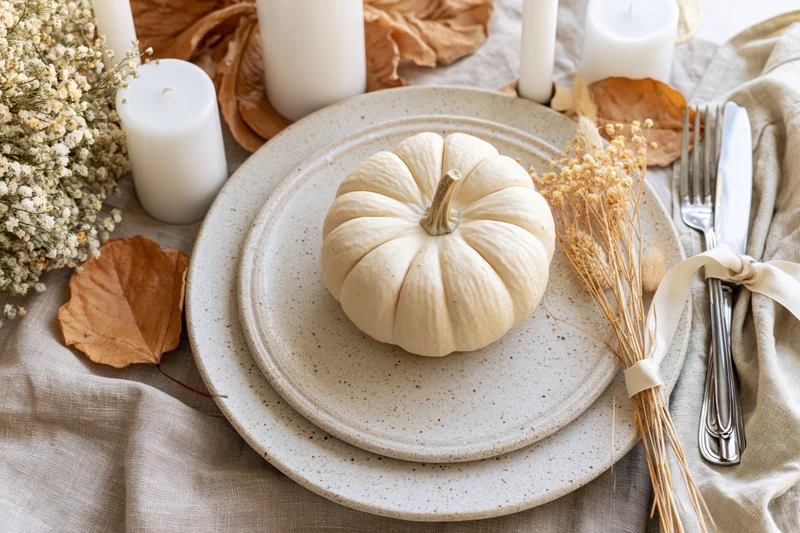 Small cream pumpkin on stacked ceramic plates at autumn table setting with candles and dried foliage