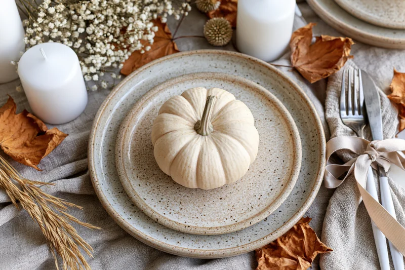 Small cream pumpkin centered on stacked ceramic plates in an autumn table setting with dried leaves, candles, and linen napkins