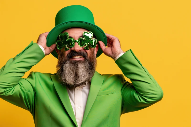 Bearded man in green suit and hat adjusting clover sunglasses against yellow background