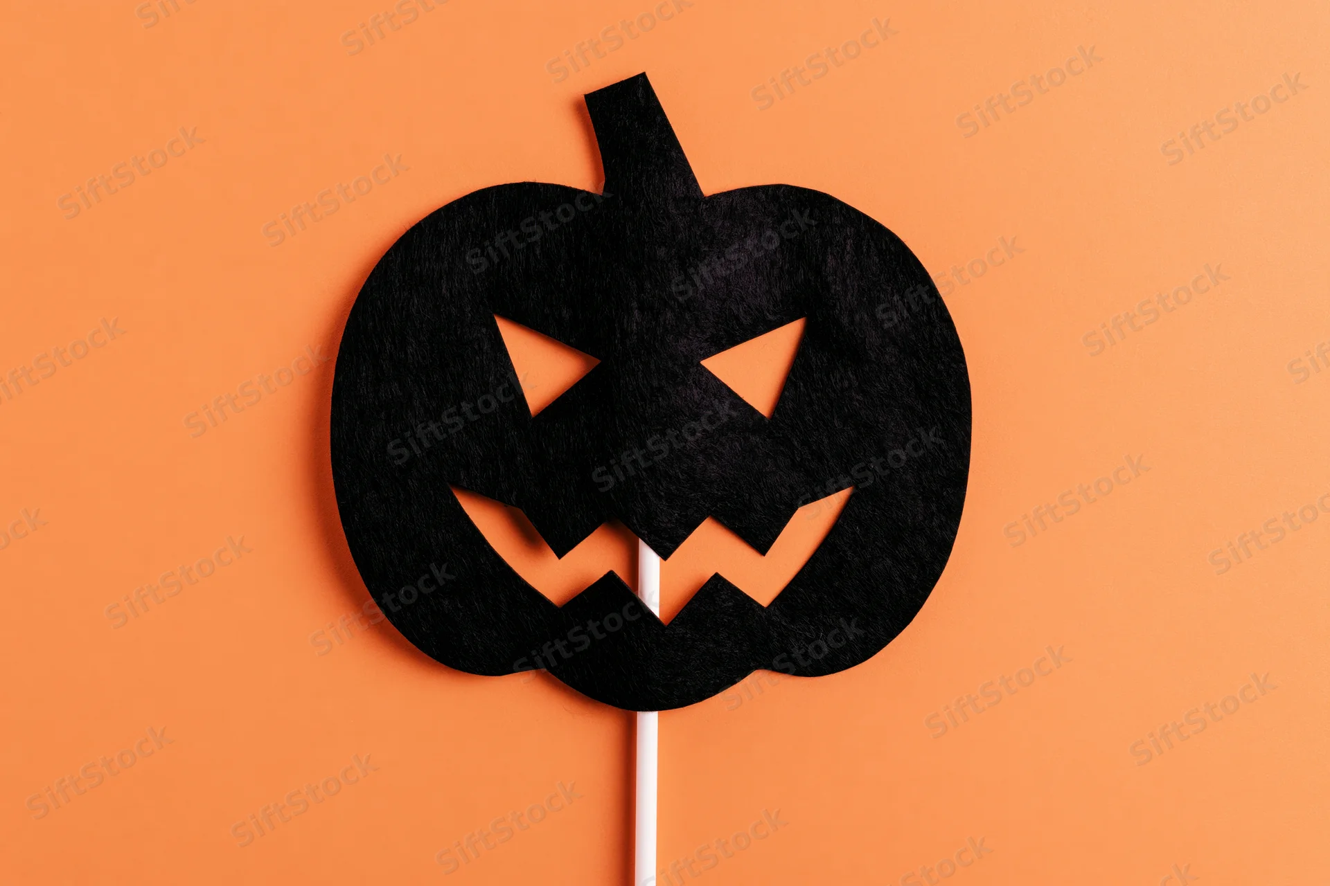 Black felt jack-o-lantern mask on white stick against orange background