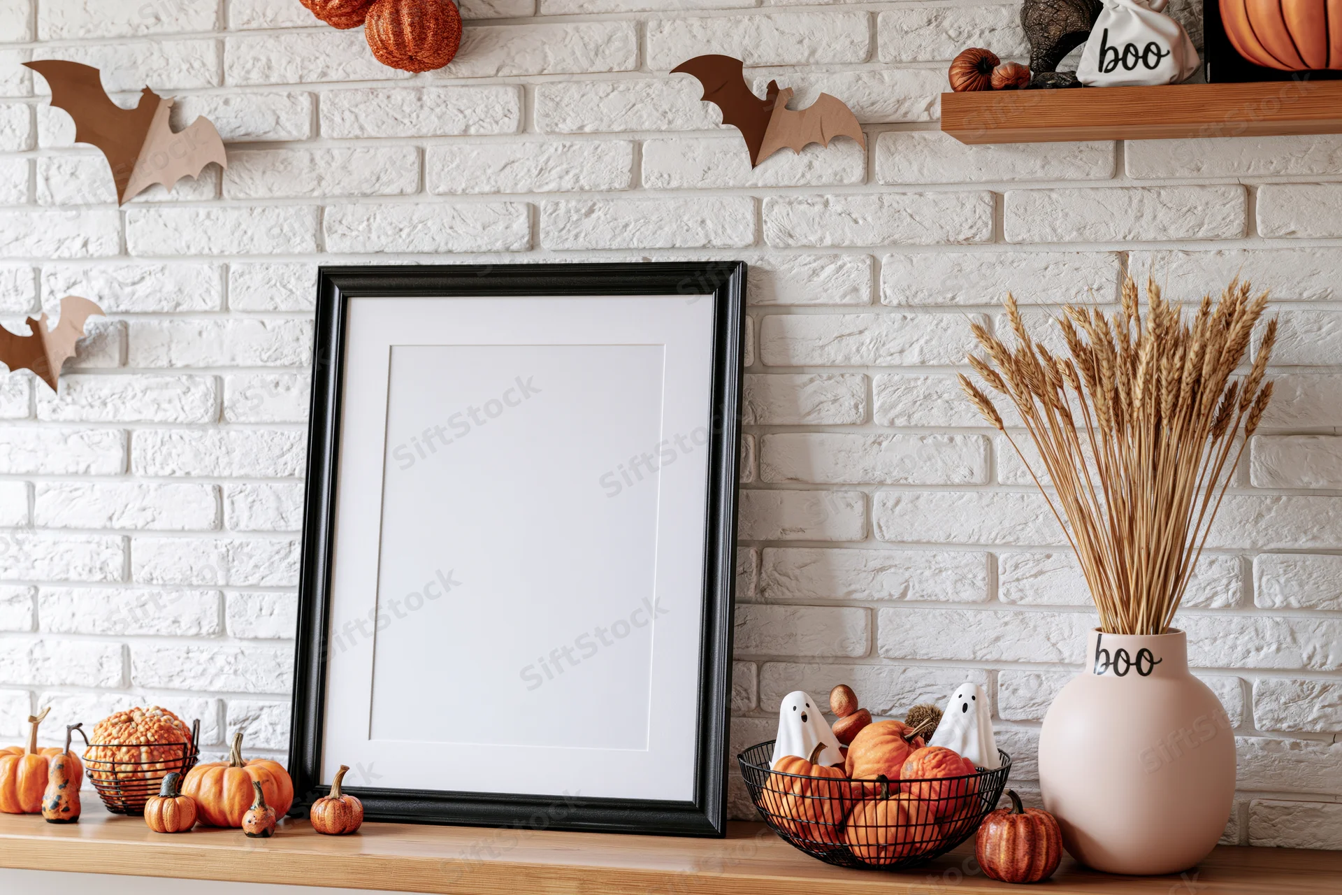 Black picture frame with white mat leaning on wooden shelf surrounded by halloween decorations including pumpkins, bats, and wheat in a vase against white brick wall