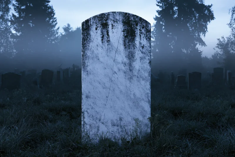 Weathered marble gravestone standing upright in overgrown cemetery at dusk surrounded by fog and trees