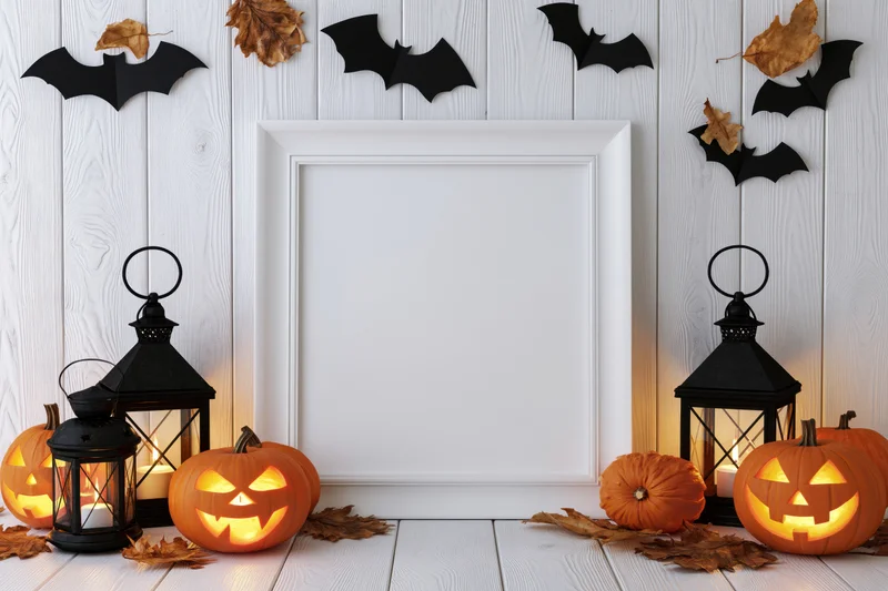 Blank white frame leaning against white wood wall surrounded by glowing jack-o-lanterns, black lanterns, paper bats and autumn leaves