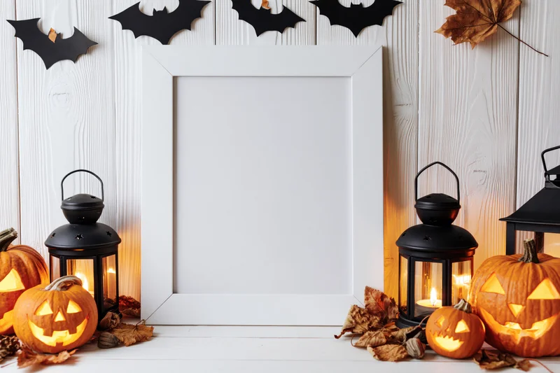 White picture frame leaning against wood wall surrounded by carved pumpkins, black lanterns, and bat decorations for halloween display