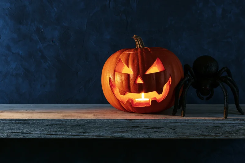 Carved jack-o-lantern glowing with candlelight beside a black spider on a wooden table against a dark textured wall
