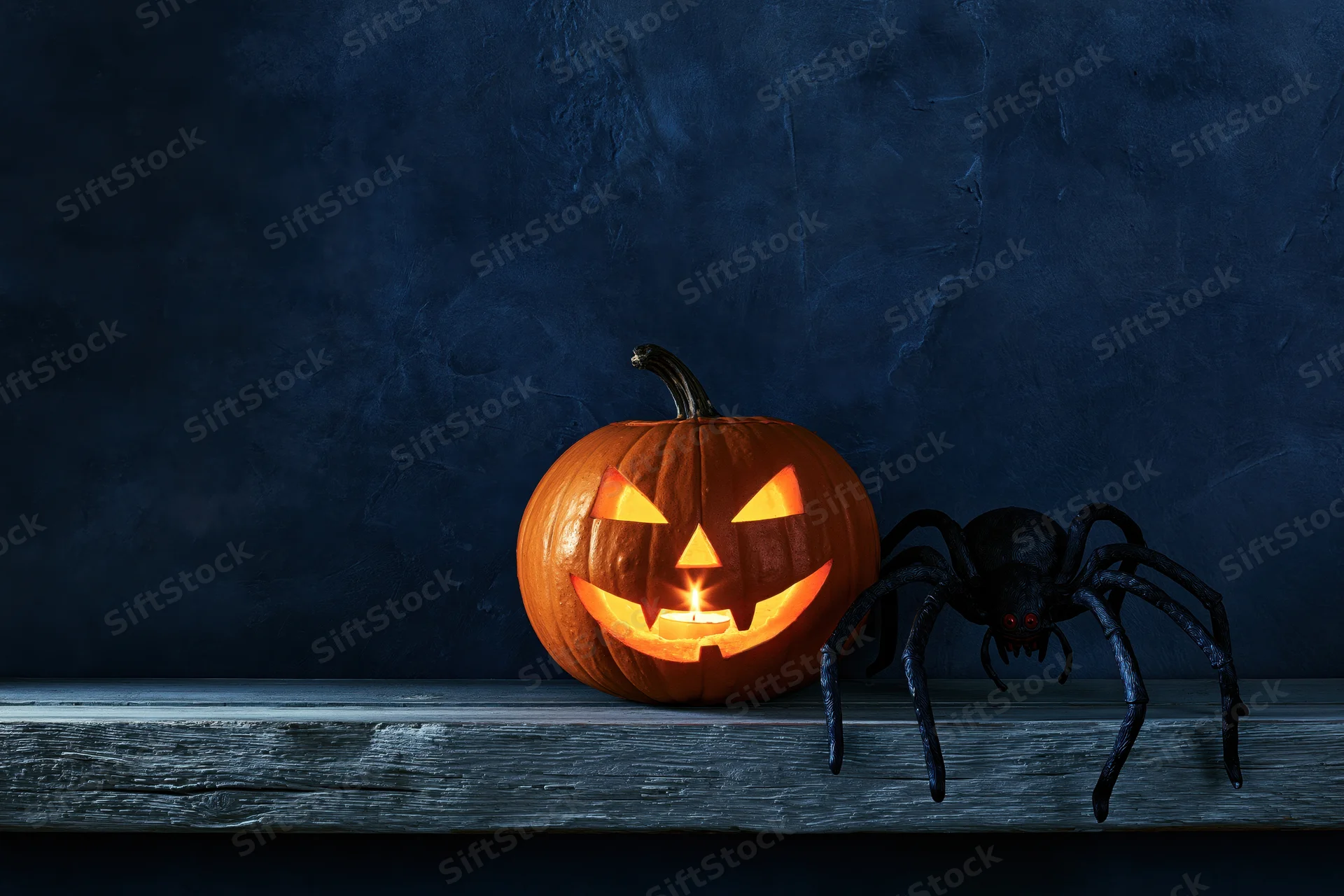 Carved jack-o-lantern glowing with candlelight beside a black spider on a wooden shelf against a dark textured wall