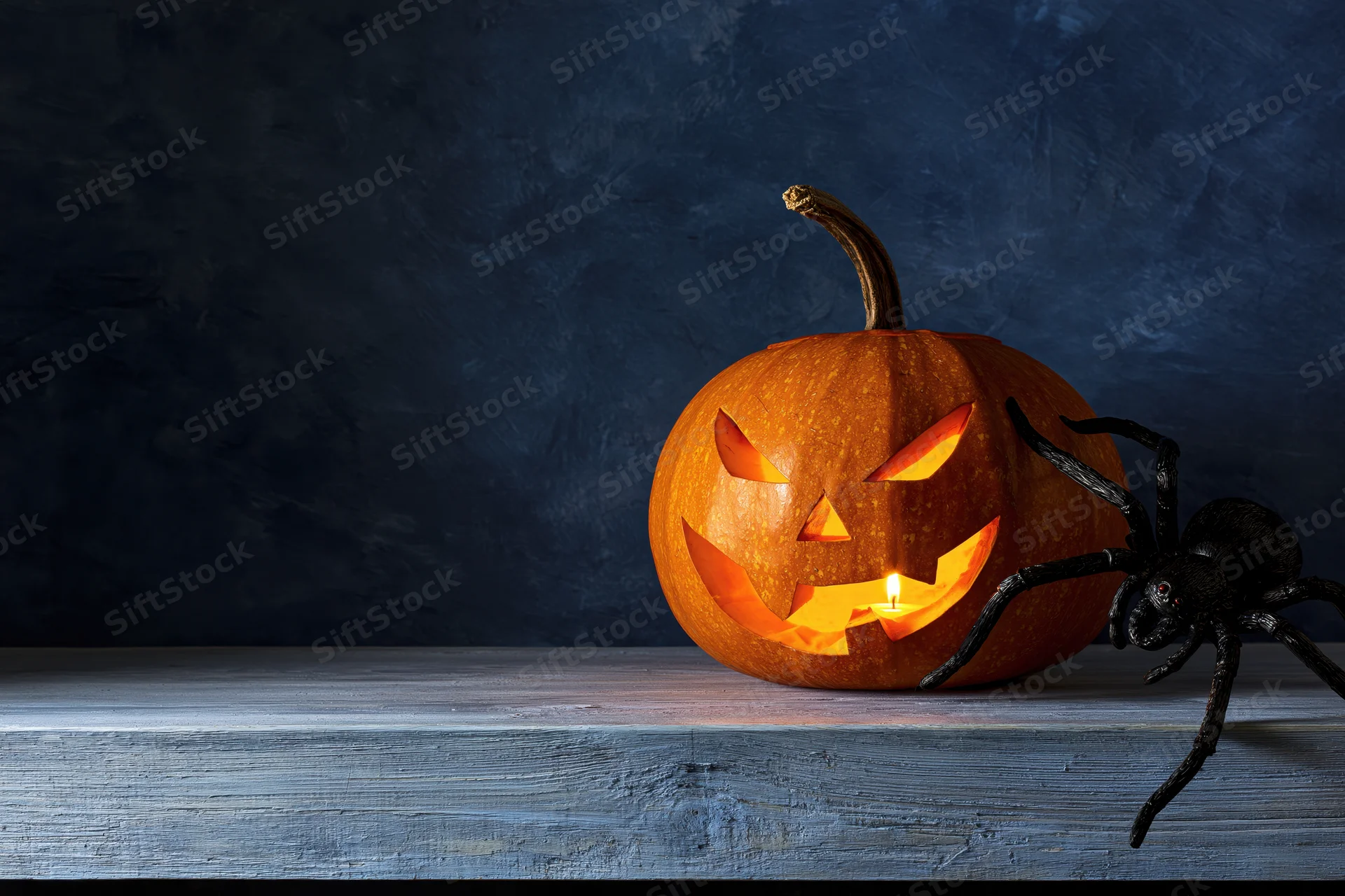 Carved jack-o-lantern glowing with candlelight beside a black spider on a wooden table against dark background
