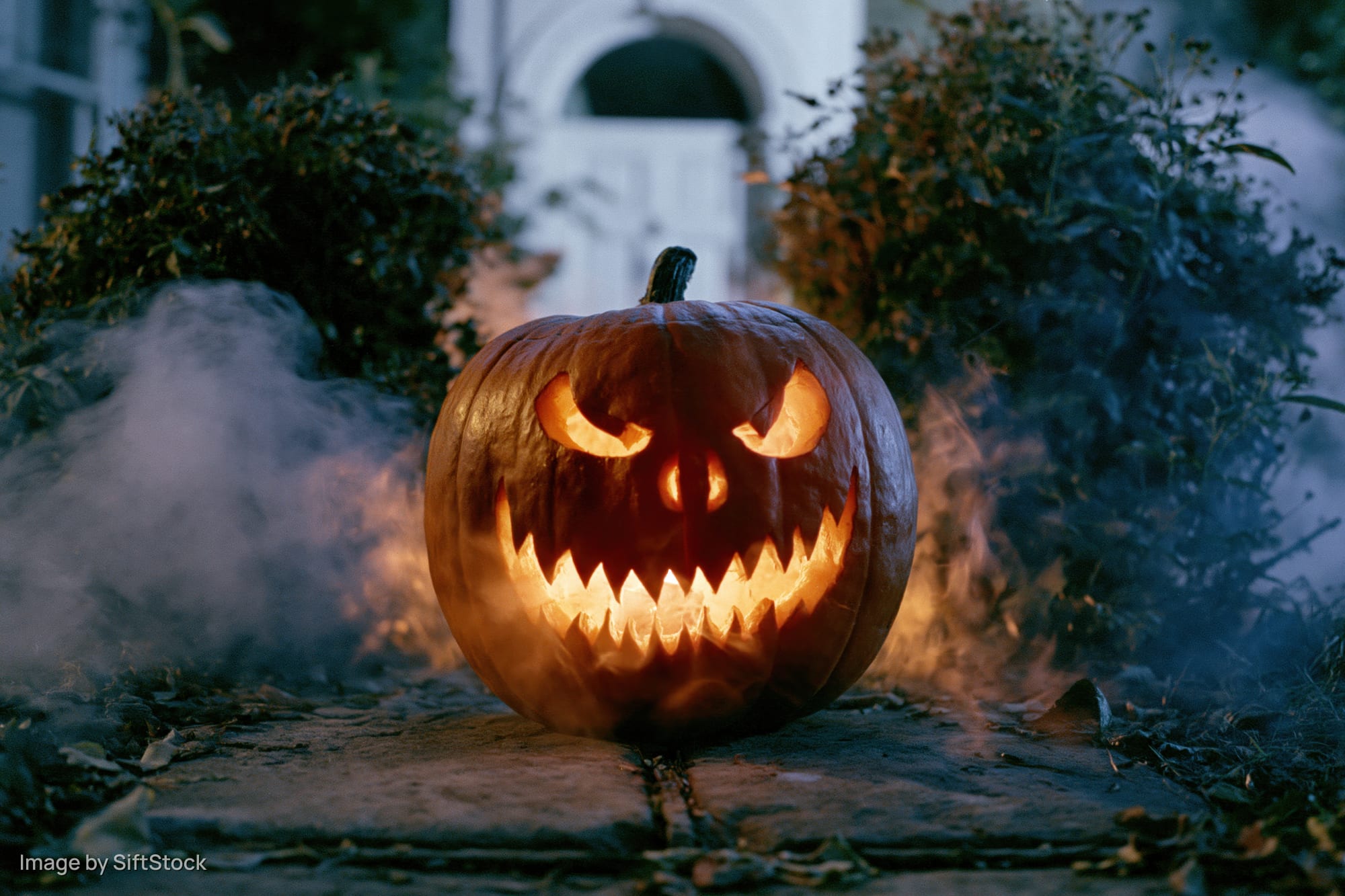 Carved jack-o-lantern with glowing evil grin on stone pathway at night surrounded by smoke and autumn foliage