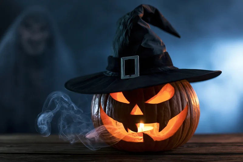 Carved jack-o-lantern with witch hat and candlelight on wooden table, smoke drifting, ghostly figure blurred in background