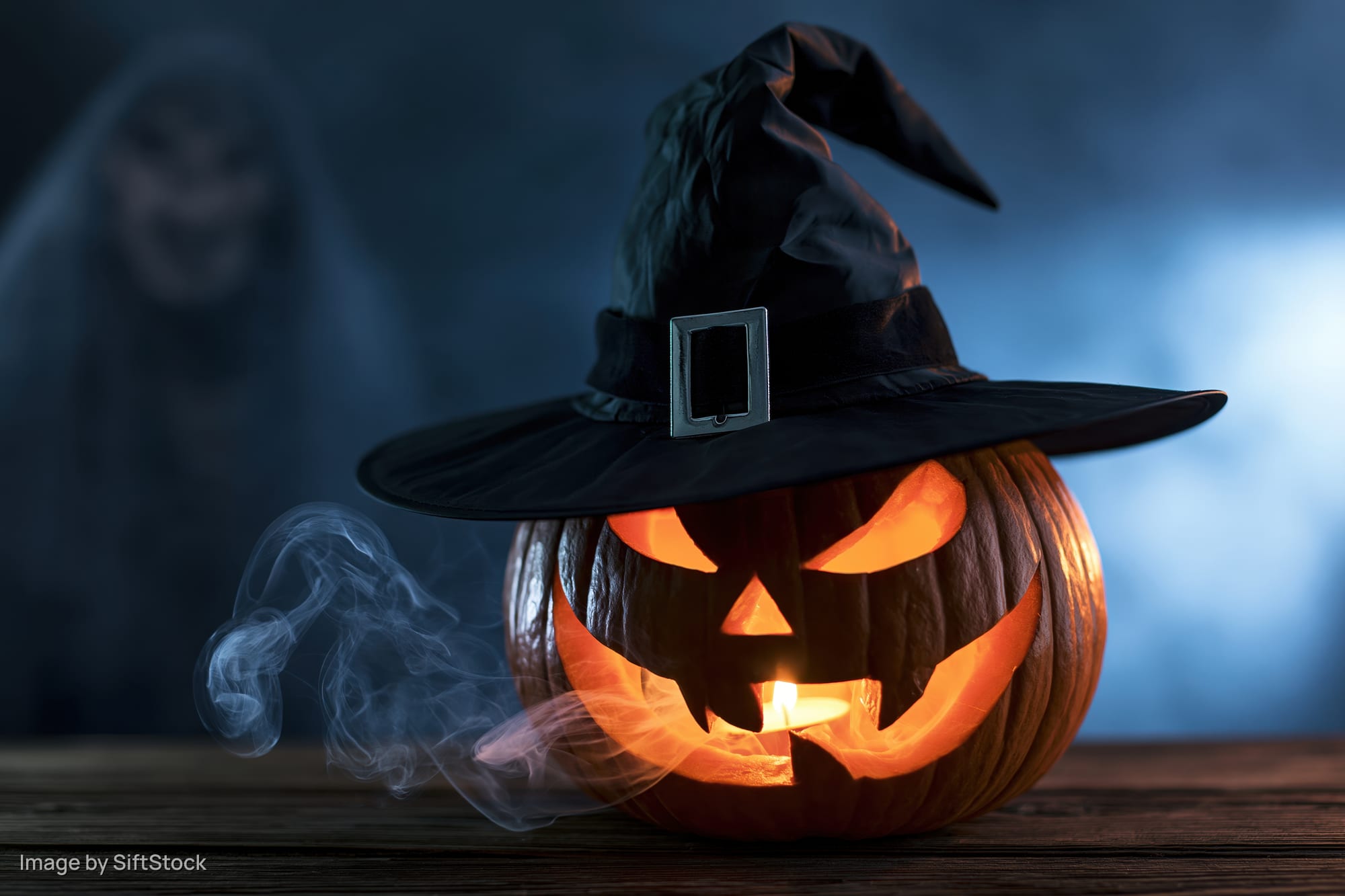 Carved jack-o-lantern with witch hat and candlelight on wooden table, smoke drifting, ghostly figure blurred in background