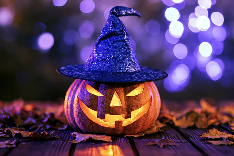 Carved jack-o-lantern with glowing face wearing blue glittery witch hat on wooden table surrounded by dry leaves at night