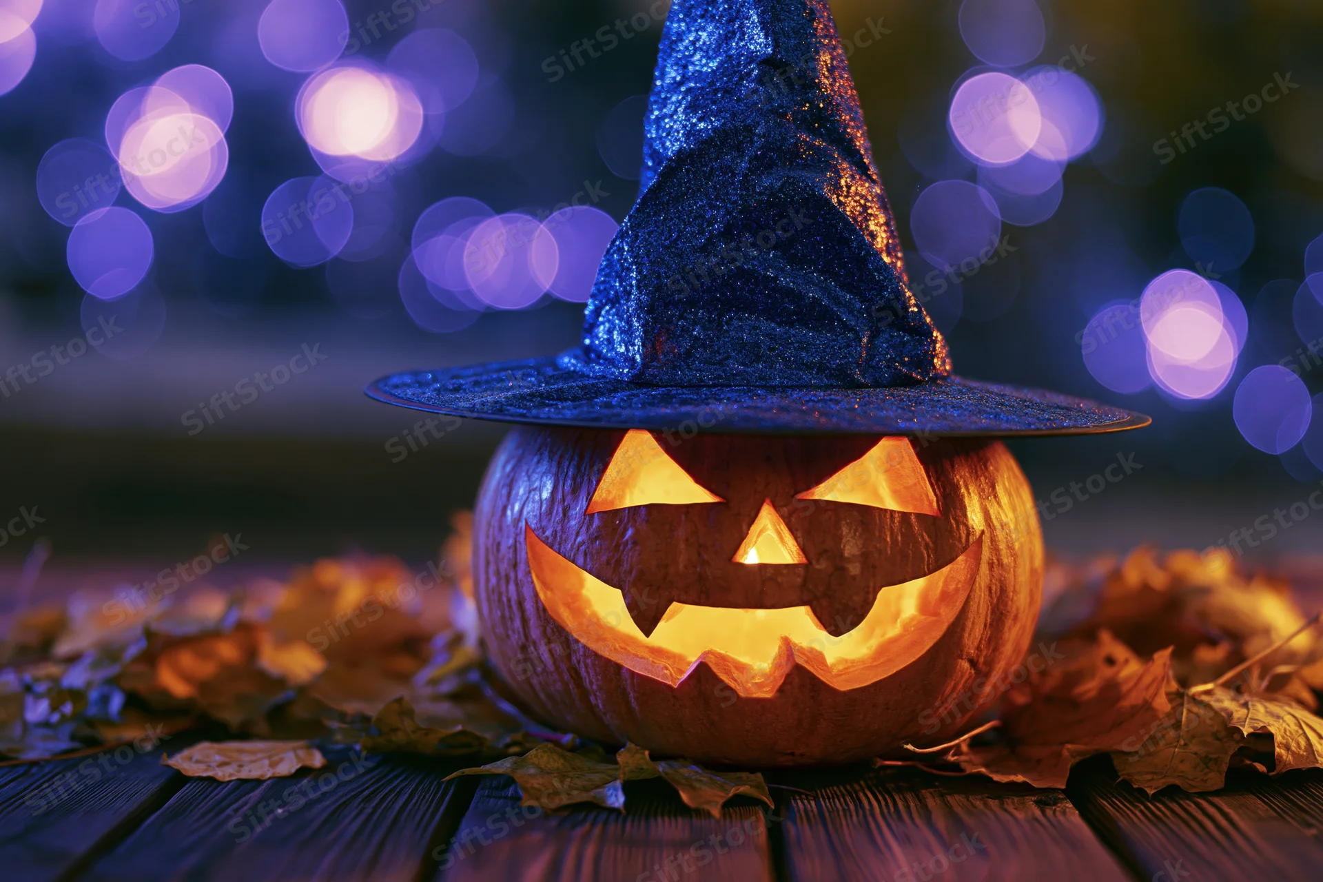 Carved jack-o-lantern with blue glittery witch hat glowing on wooden table surrounded by autumn leaves at night