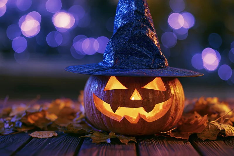 Carved jack-o-lantern with blue glittery witch hat glowing on wooden table surrounded by autumn leaves at night