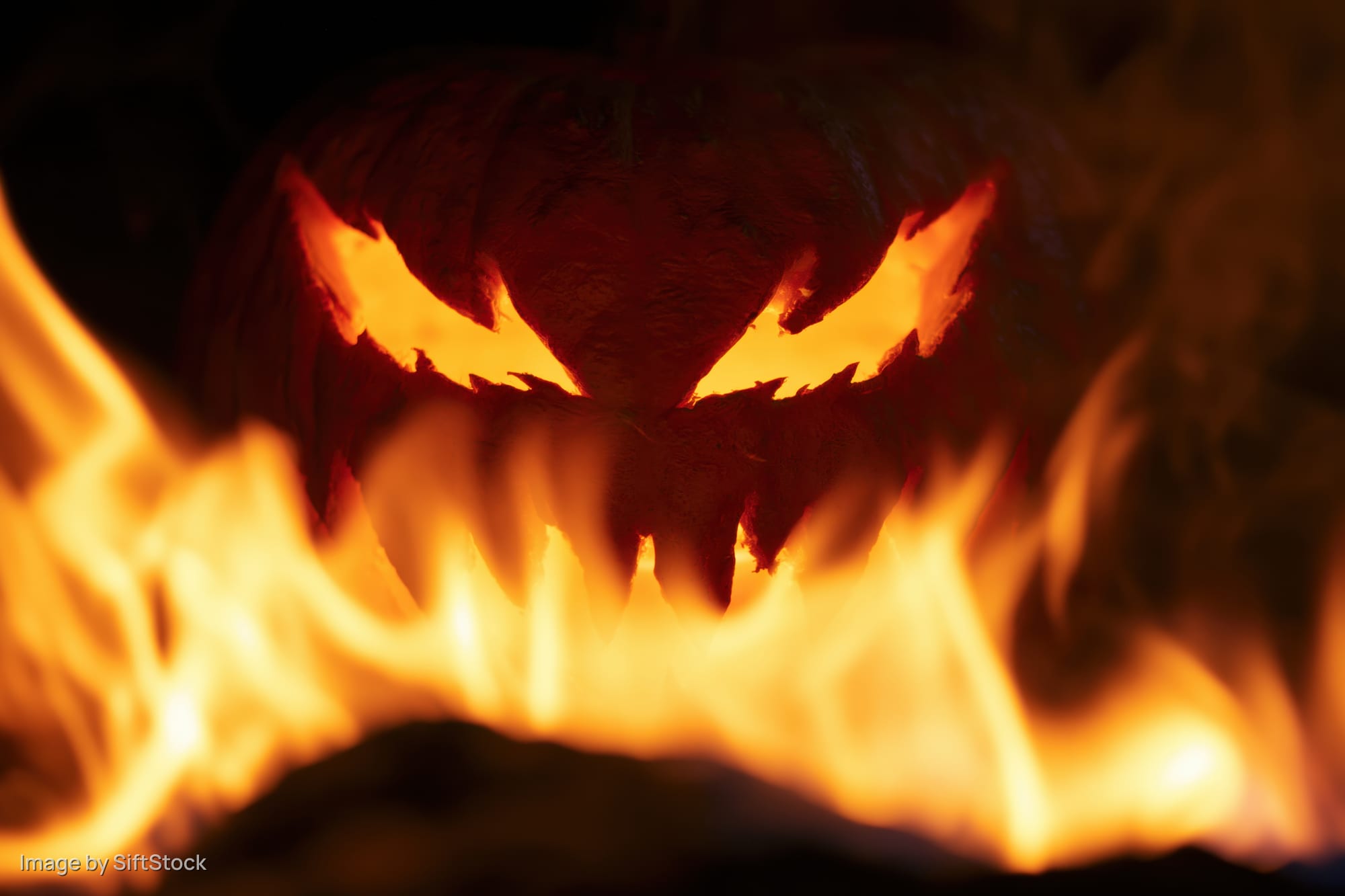 Carved jack-o-lantern with jagged eyes glowing behind flames at night