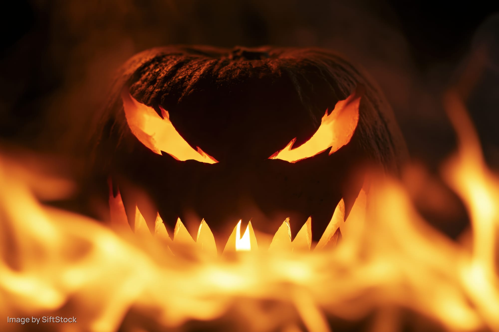 Carved jack-o-lantern with jagged glowing eyes and sharp teeth surrounded by flames at night