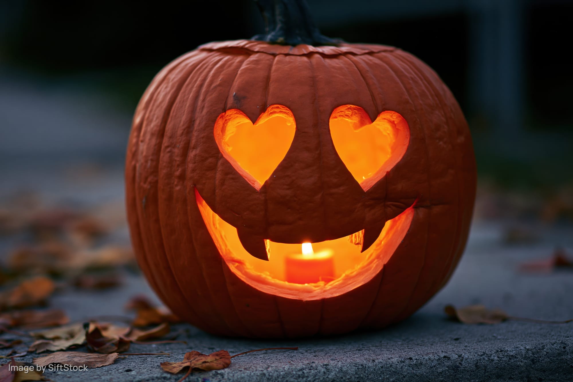 Carved pumpkin with heart-shaped eyes and smiling mouth lit by candle on ground surrounded by fallen leaves at night