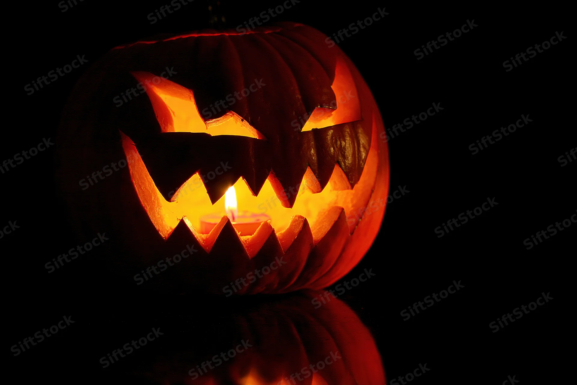 Carved pumpkin with jagged grin illuminated by candlelight on dark reflective surface