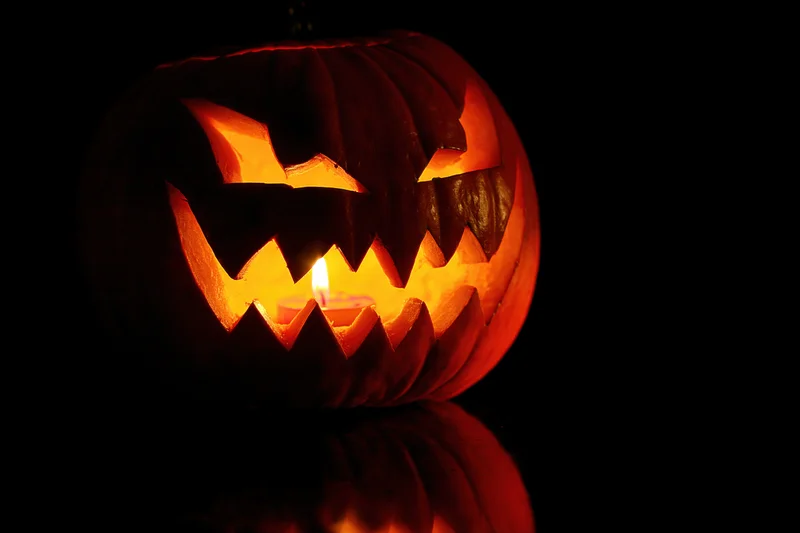 Carved pumpkin with jagged grin illuminated by candlelight on dark reflective surface