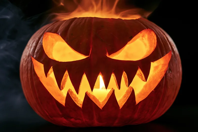 Carved pumpkin with jagged teeth and glowing eyes lit by candle in dark smoky setting