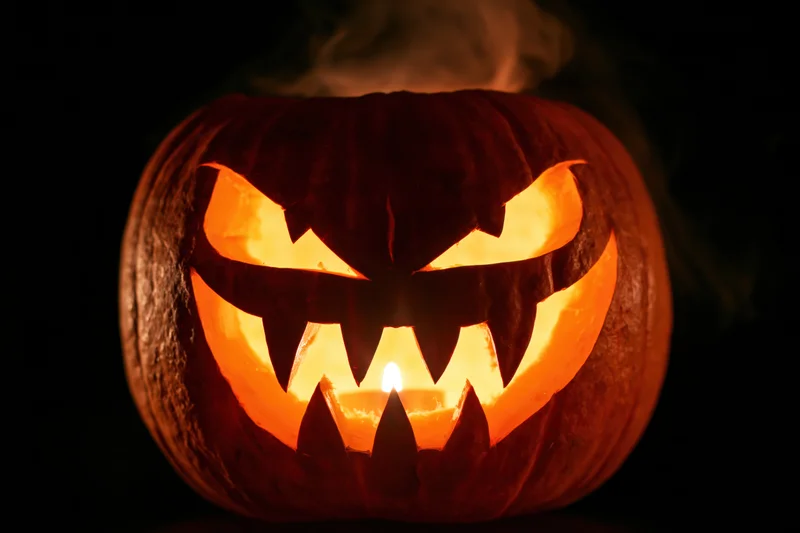 Carved pumpkin with jagged teeth and glowing eyes lit by candle in dark setting
