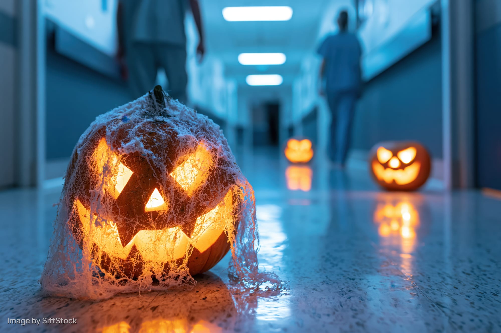 Carved glowing jack-o-lantern covered in fake cobwebs on hospital hallway floor with staff walking in background