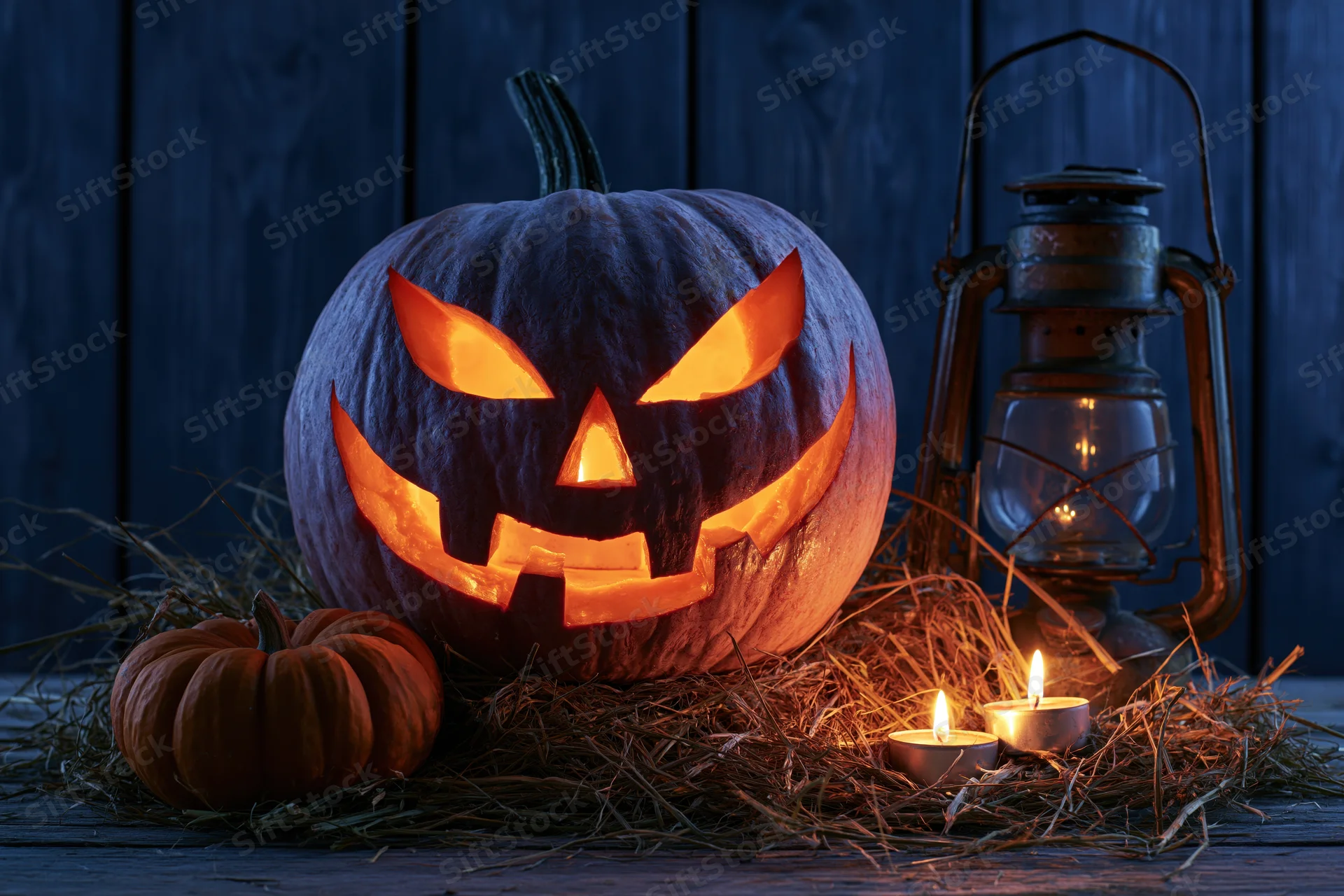 Carved jack-o-lantern glowing with candlelight on hay beside a lantern on a wooden surface at night