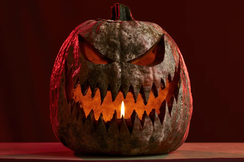 Carved jack-o-lantern with menacing toothy grin and triangular eyes lit by candle on dark red background