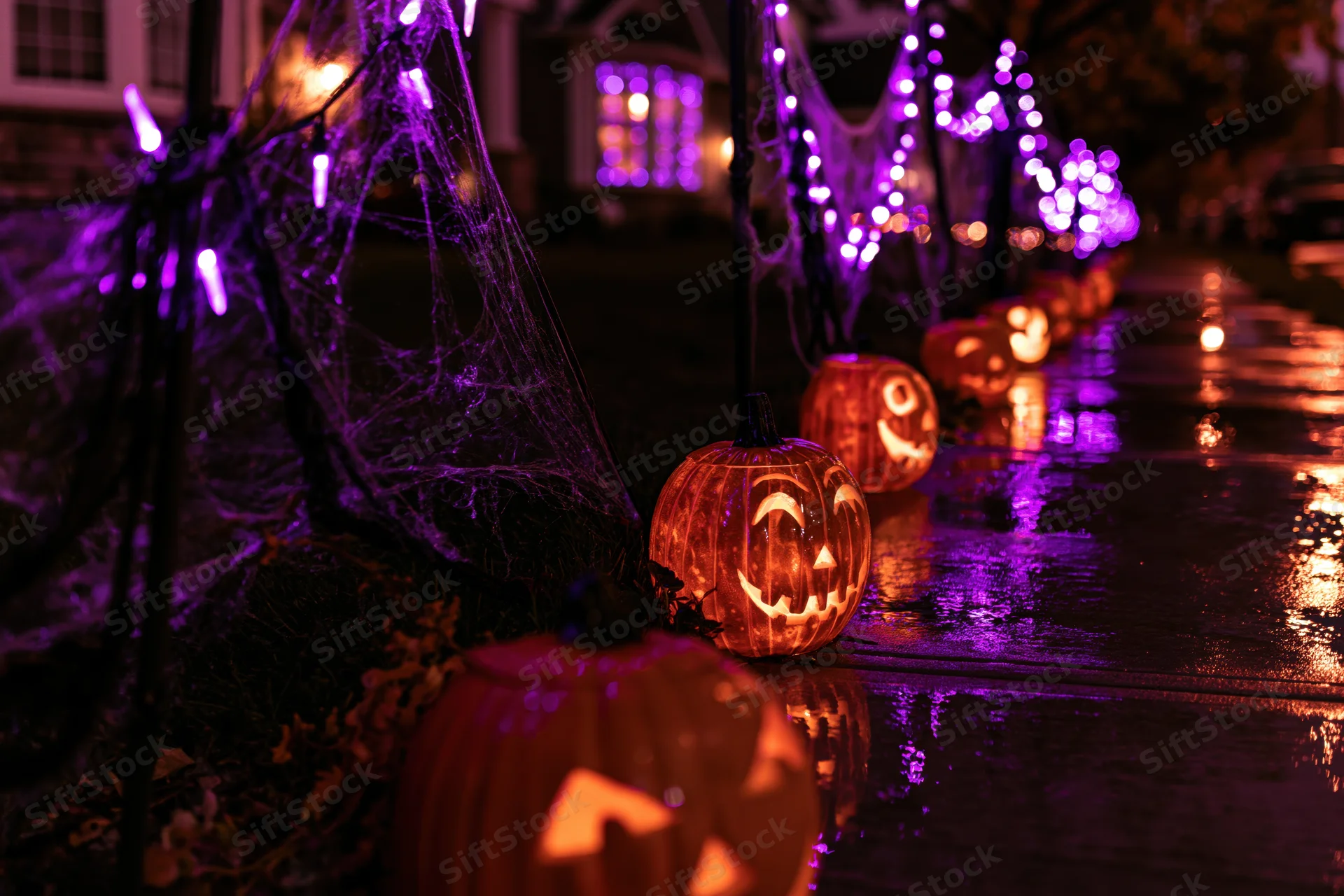 Carved jack-o-lanterns glowing orange along a wet suburban sidewalk at night with purple string lights and spiderwebs