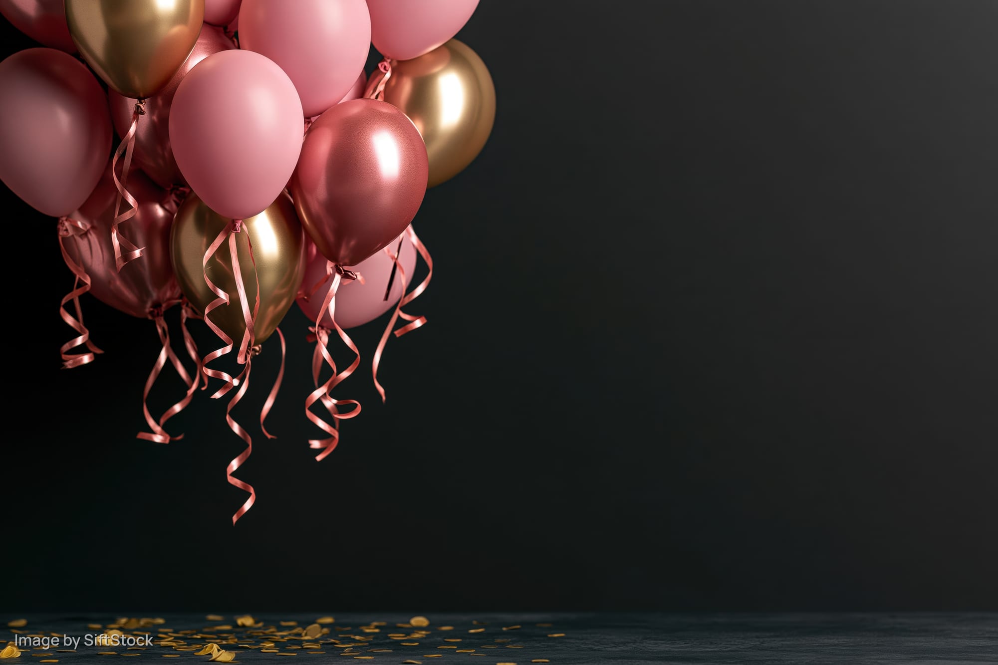 Cluster of pink and gold metallic balloons with curling ribbons floating above scattered confetti on dark floor