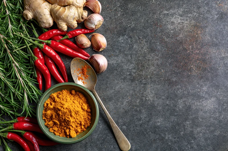 Turmeric powder in a bowl with fresh chili, garlic, ginger, and rosemary arranged along the left edge of a dark textured surface