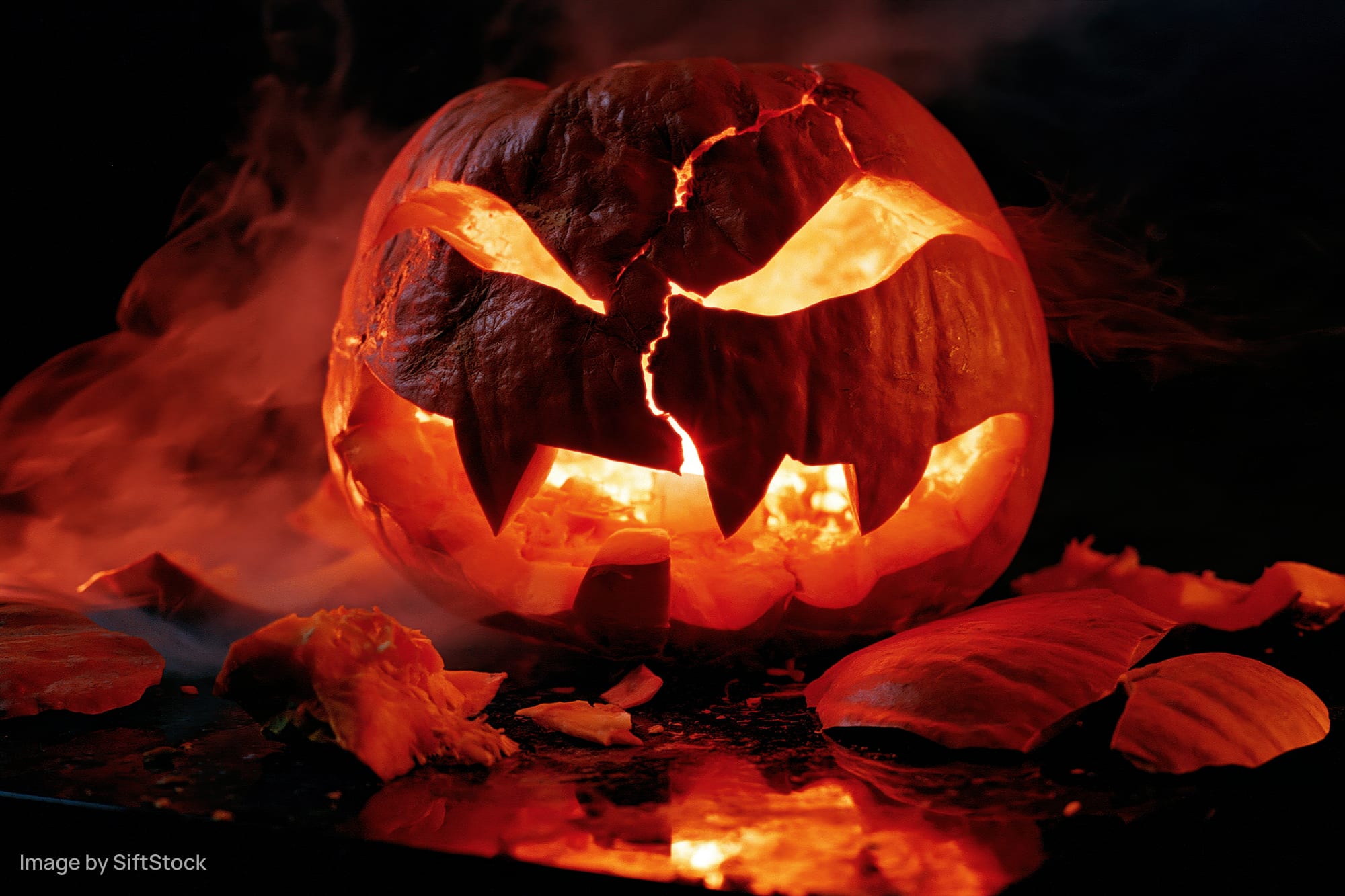 Cracked jack-o-lantern glowing with smoke and broken pieces around it on dark surface
