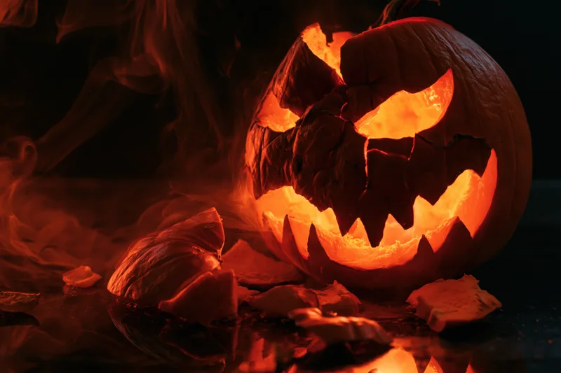 Cracked jack-o-lantern glowing with orange light on dark wet surface surrounded by broken pieces and smoke